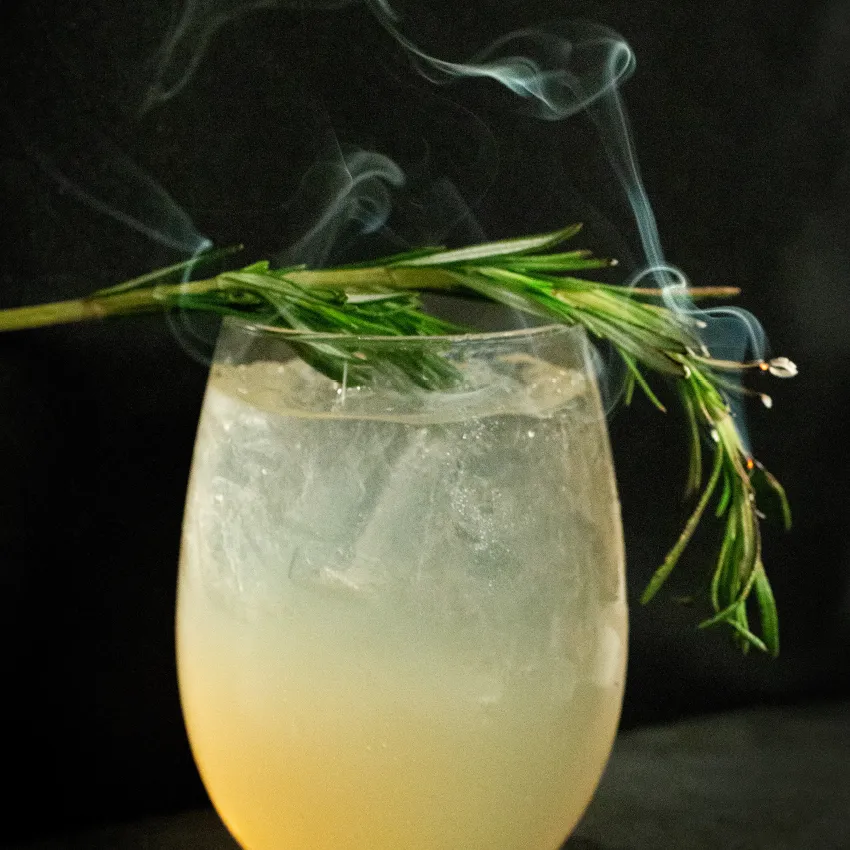 A light yellow cocktail in a wine glass with a smoking sprig of rosemary